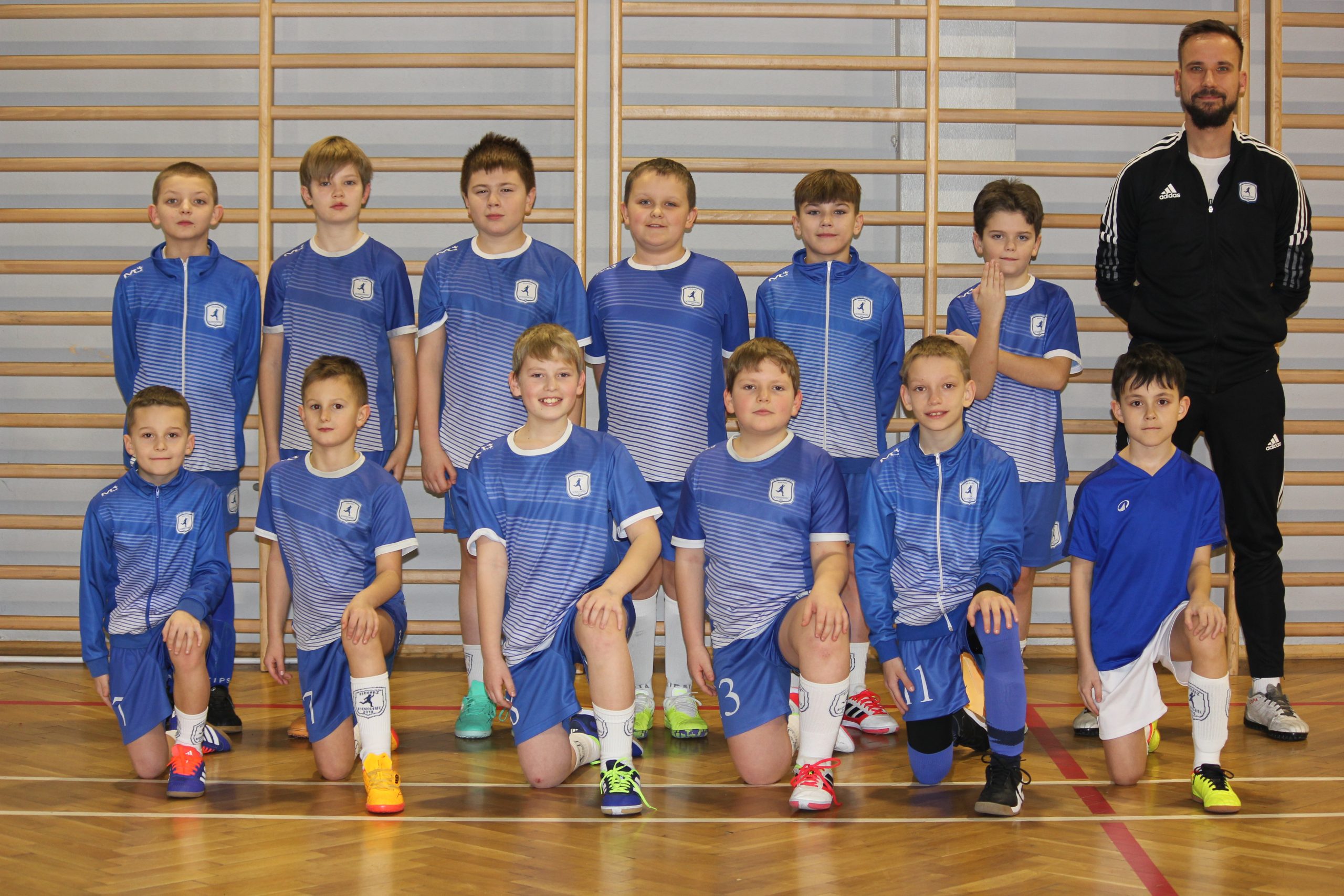 Zespół młodych chłopców w strojach sportowych oraz trener, ustawieni w szeregu w hali sportowej.